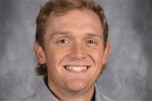 Head-and-shoulders portrait of a smiling man with blond hair against a gray studio backdrop.”