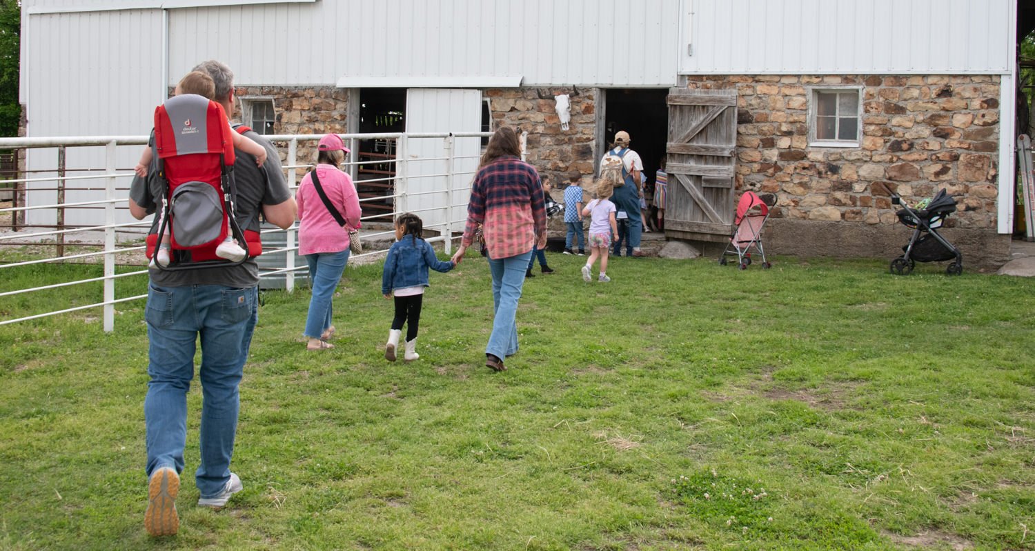 Family and visitors walk toward a rustic stone barn with white doors on a grassy farm yard, a stroller nearby.