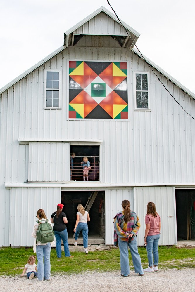 White barn with a colorful quilt-style window; group of people gathered outside, a child on a balcony.