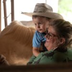 Woman holding a smiling toddler in a straw hat, inside a barn near a horse.