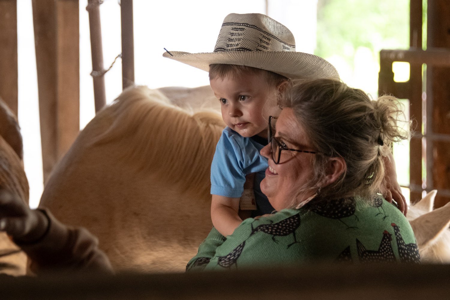 Woman holding a smiling toddler in a straw hat, inside a barn near a horse.