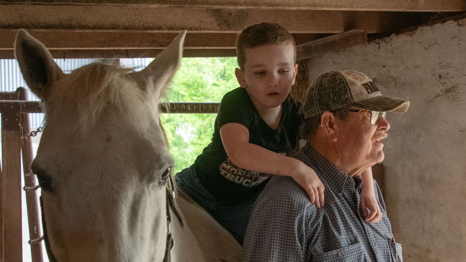 Boy on a man's shoulders beside a white horse in a barn/stable.