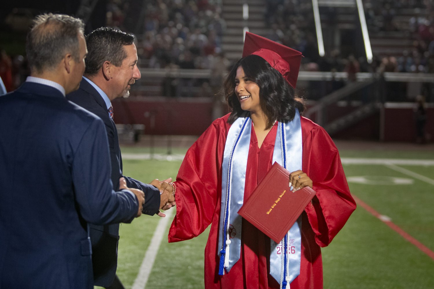 Graduate in a red cap and gown shakes hands with a man in a suit on a football field at night, holding a red diploma cover in her other hand.