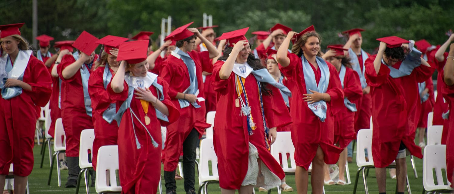Group of graduates in red gowns and caps outdoors, adjusting their caps as they walk past white chairs.