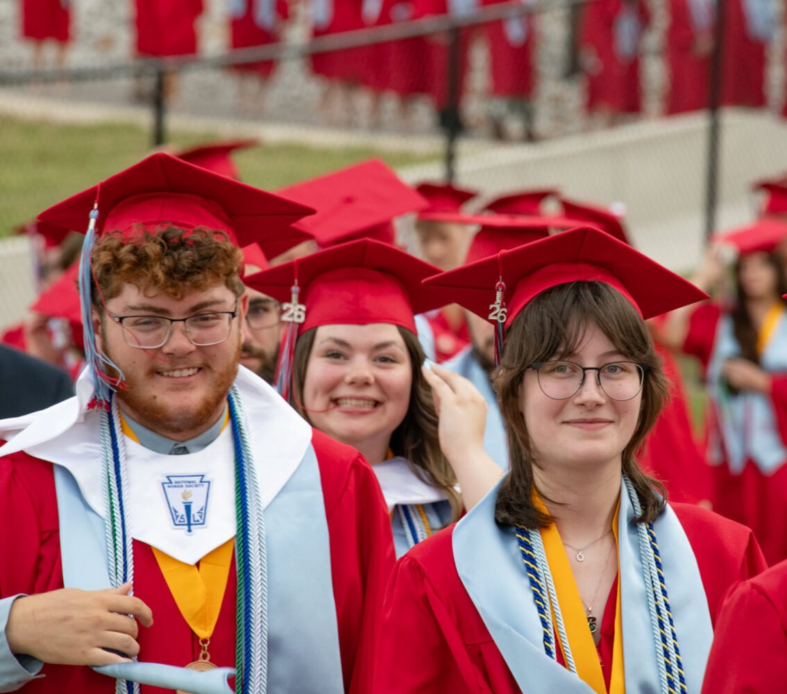 Three graduates in red caps and gowns pose for a photo at a commencement, smiling with cords and a stole visible.