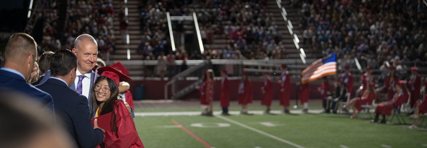A graduate in a red cap and gown smiles while shaking hands with adults on a football field during a ceremony, fellow graduates and a marching band in the background.
