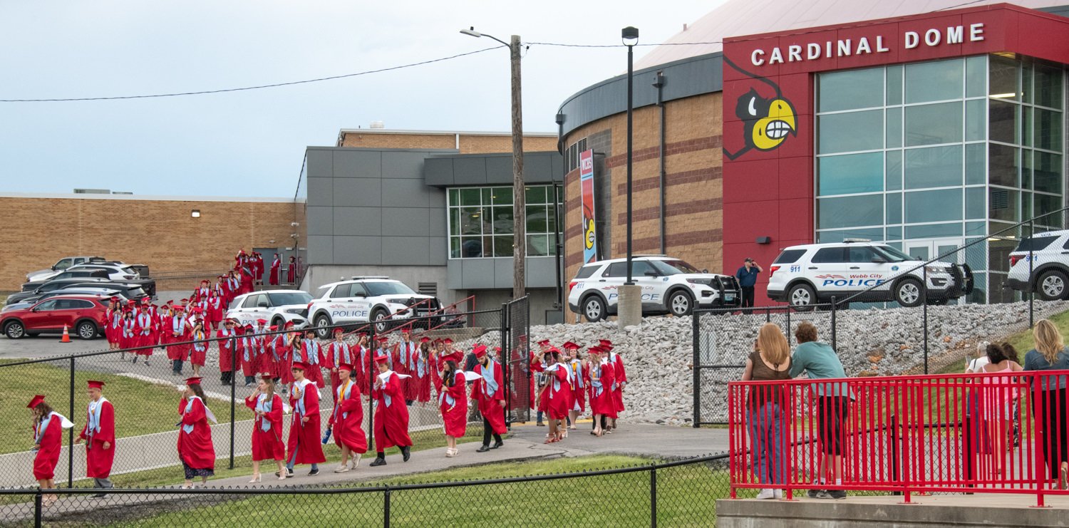Graduates in red gowns walk in a procession outside Cardinal Dome, with bystanders behind a red railing.