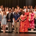 Group of students on a stage posing for a photo after a performance, dressed in formal and semi-formal attire on risers.