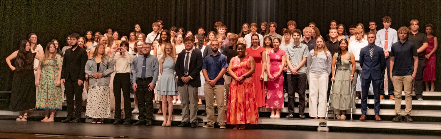 Group of students on a stage posing for a photo after a performance, dressed in formal and semi-formal attire on risers.