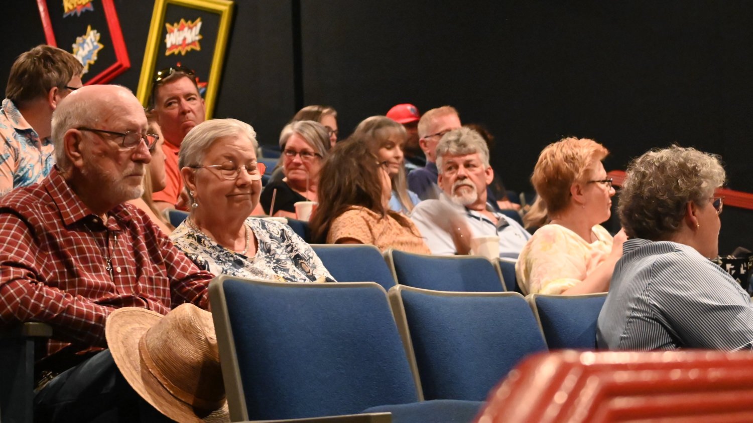Audience members seated in a dark theater, foreground shows an elderly couple smiling while others chat and watch a performance.