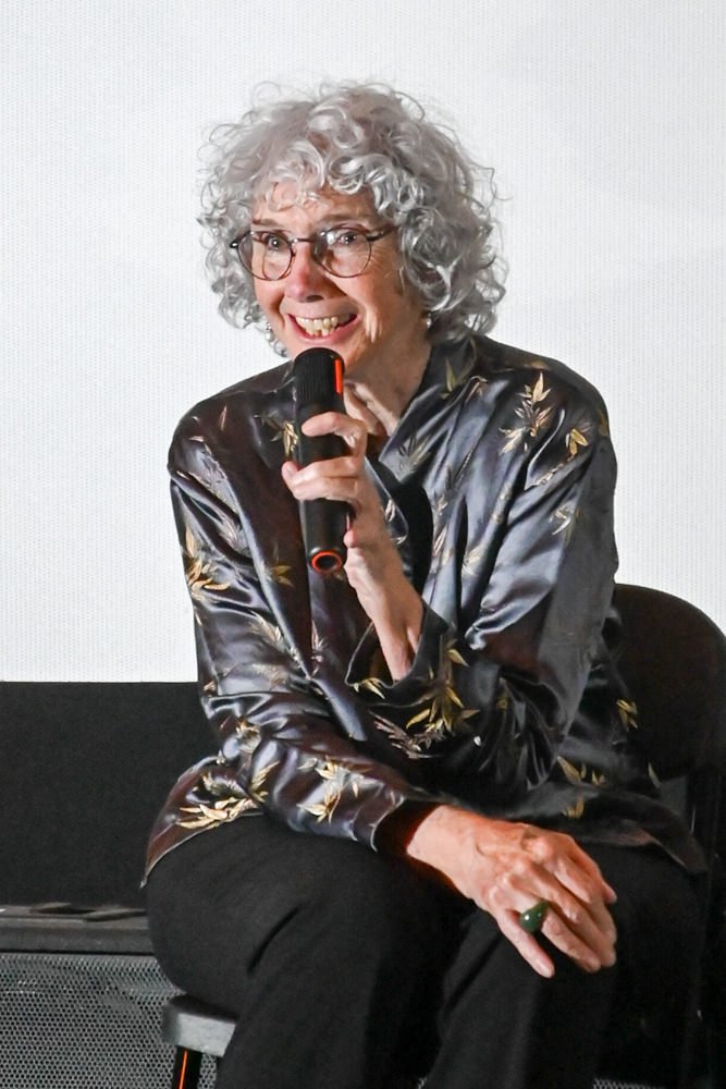 Older woman with curly gray hair and glasses, holding a microphone and smiling during a talk.