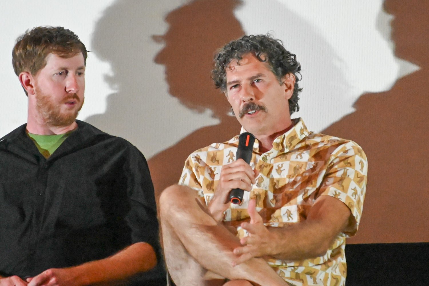 Two men seated on a stage; the man on the right speaks into a handheld microphone while the other listens.
