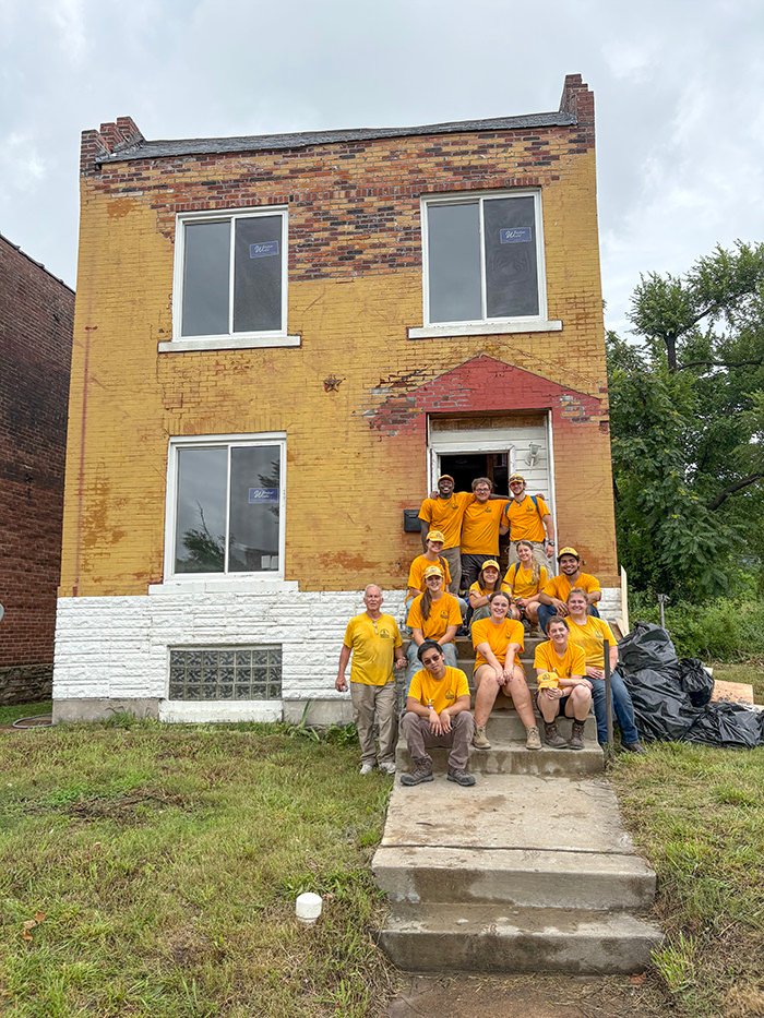 Group of volunteers in yellow shirts posing on front steps of a brick house under renovation.