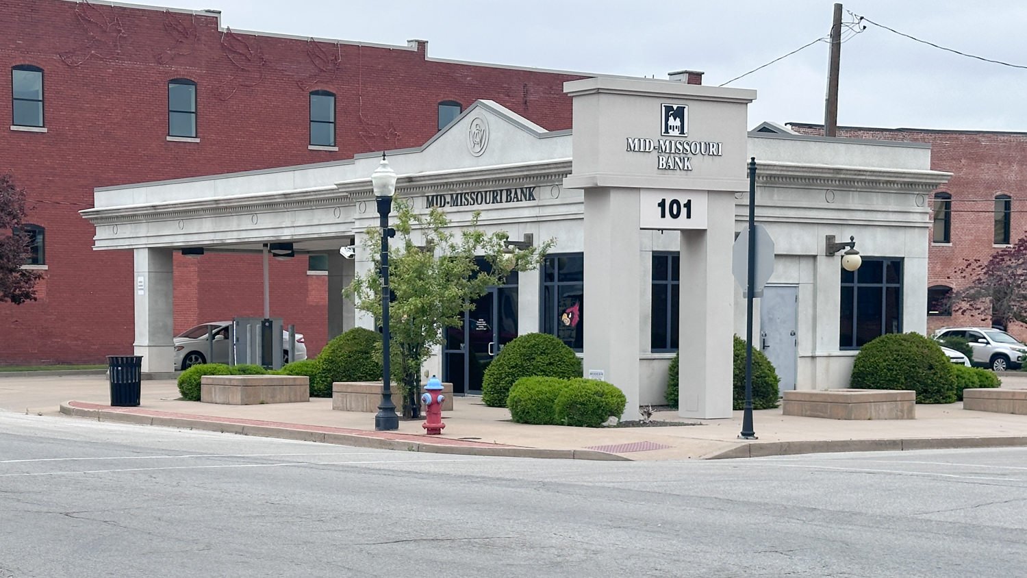 Mid-Missouri Bank on a street corner with a beige façade and a 101 pillar, surrounded by trimmed bushes and a red fire hydrant in front.