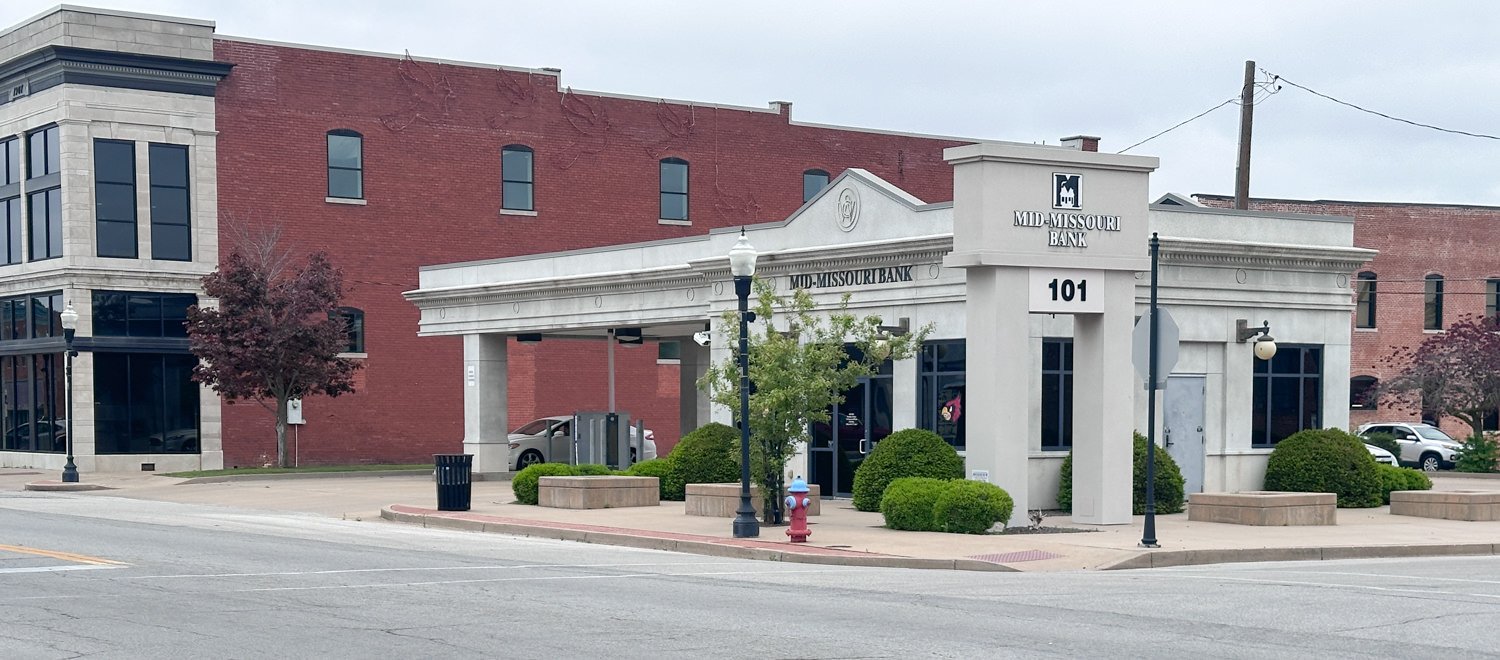 Corner view of a Mid-Missouri Bank with a white stone facade, the number 101, and trimmed bushes on the sidewalk under a cloudy sky.