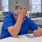 Man in a blue shirt sits at a round table, head in hand, writing on a form with a pen.