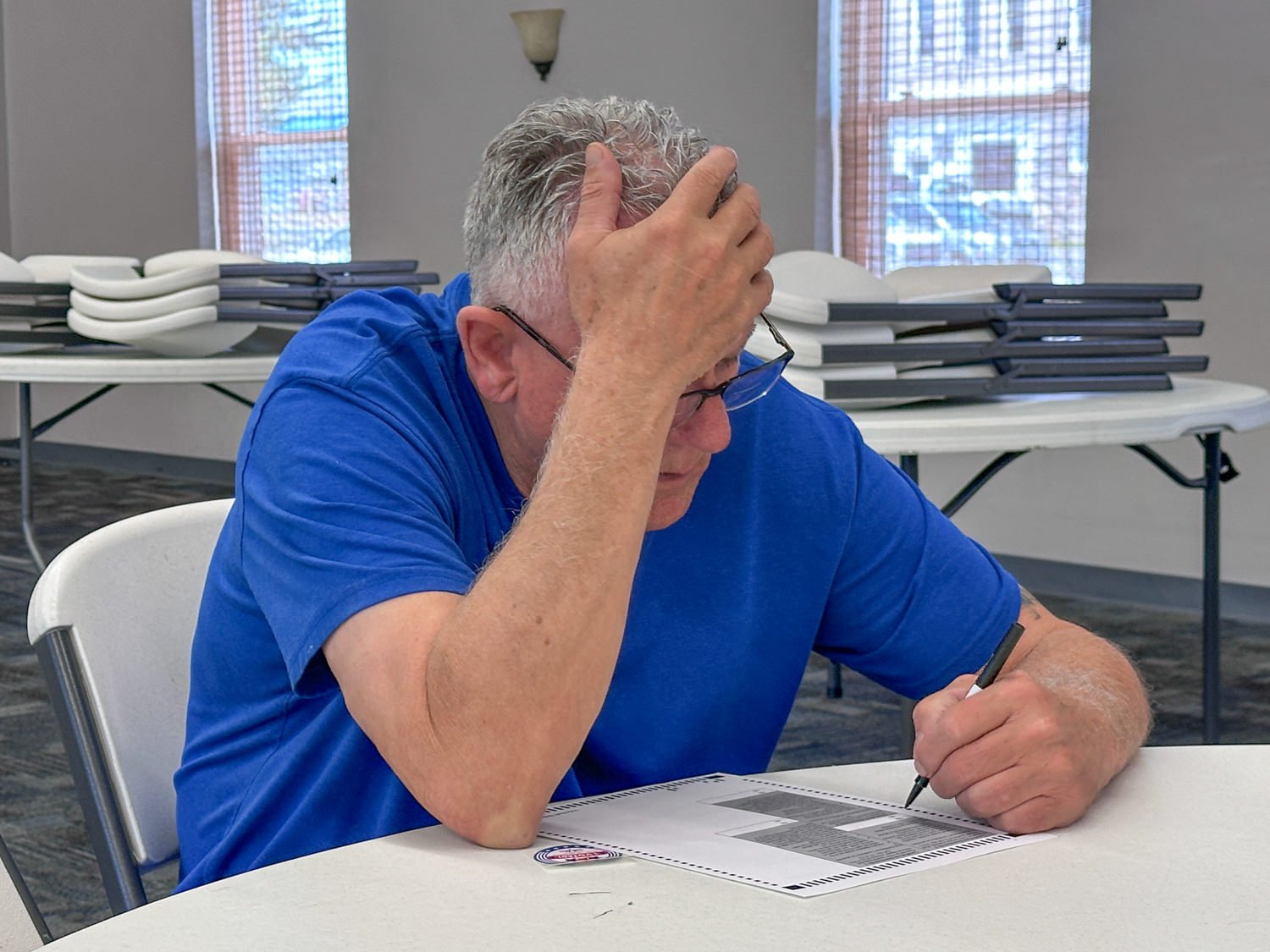 Man in a blue shirt sits at a round table, head in hand, writing on a form with a pen.