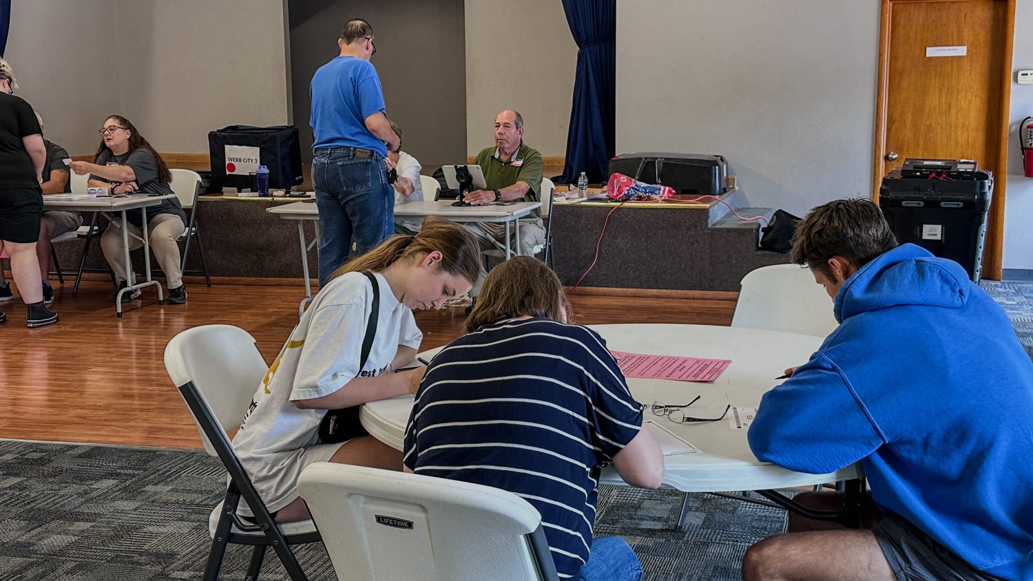 People sit at round tables writing on pink forms in a community center or election site; volunteers work at a back table in the background.