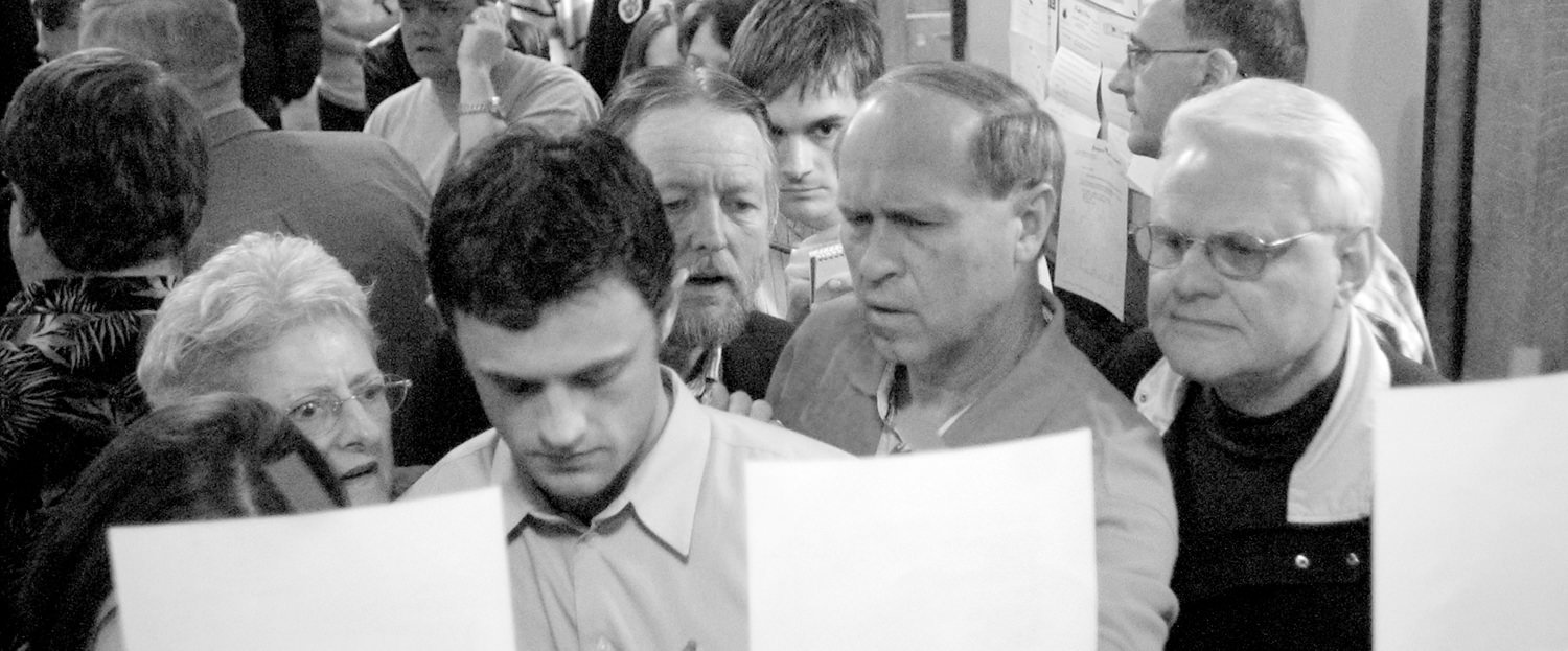 Crowded indoor scene of adults closely examining large white posters held up by participants at an event.