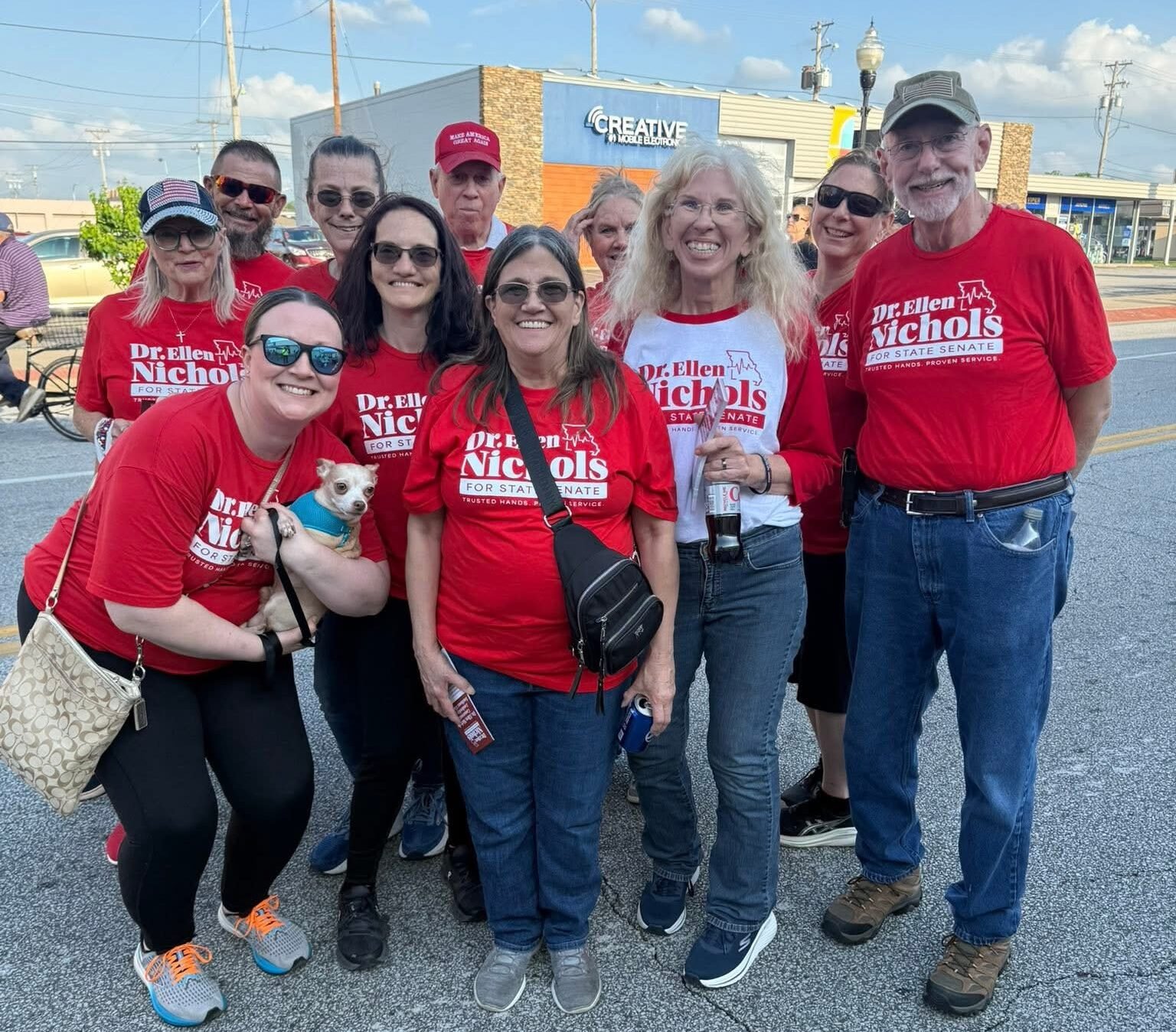 Group of volunteers in red 'Dr. Ellen Nichols for State Senate' shirts pose together outside a storefront. Candid campaign photo with a small dog in front left.