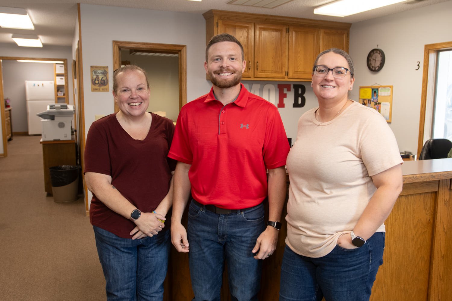 Three smiling adults stand side by side in a reception area with wooden cabinets in the background. Center is a man in a red polo; flanking him are two women in casual tops.