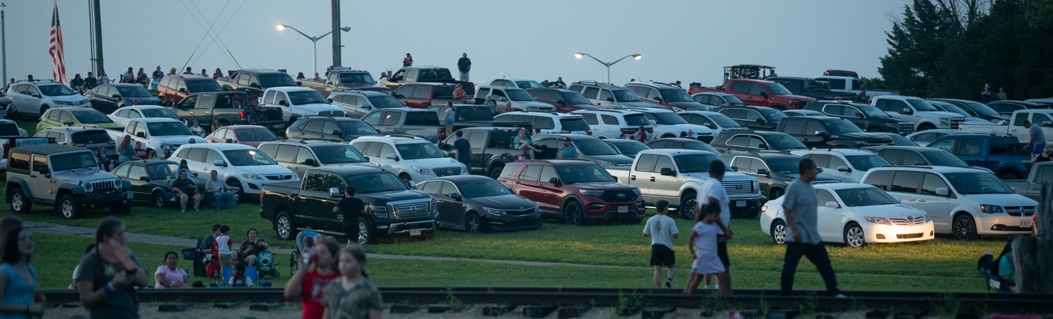 A crowded outdoor parking lot at dusk with hundreds of parked cars and groups of people walking along the grassy edge.