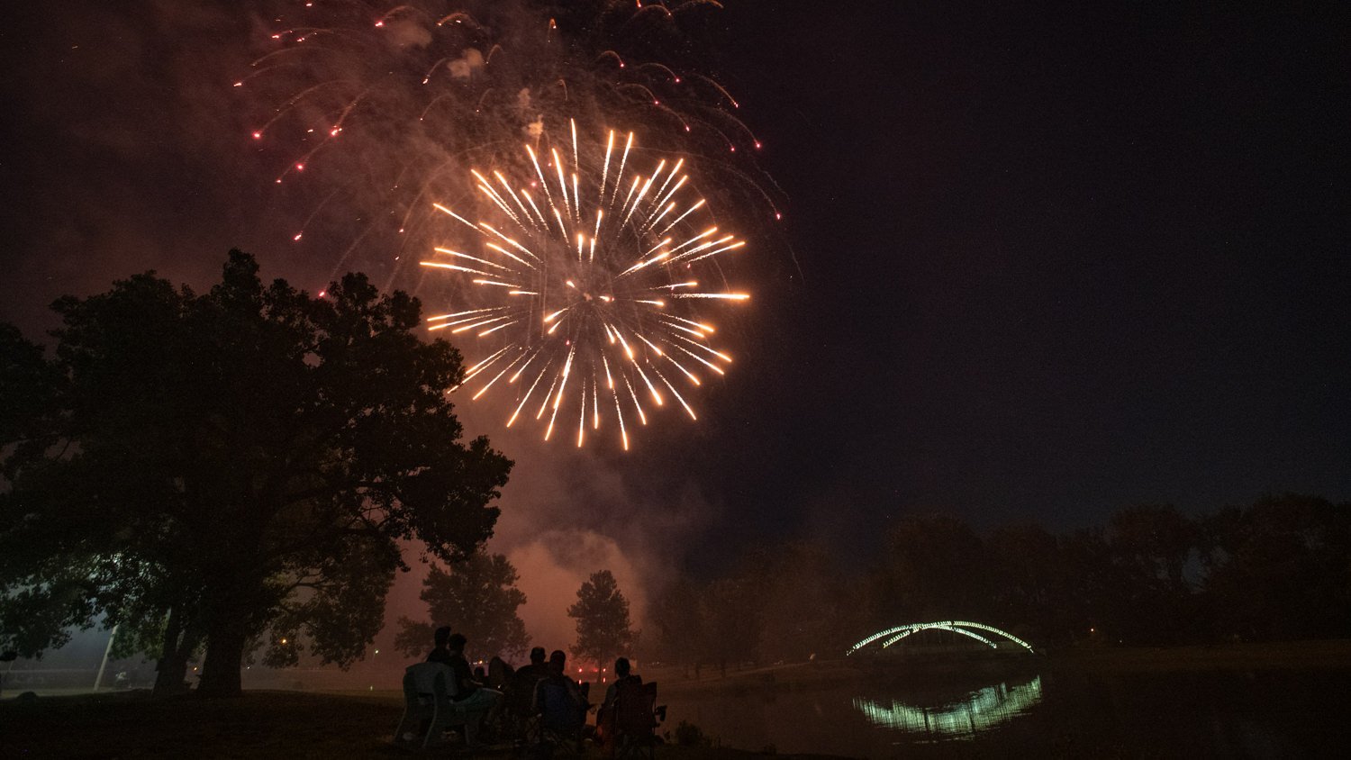 Fireworks over a park lake at night; silhouettes of trees and people on chairs watch from the shore, an illuminated arch bridge reflected in the water.