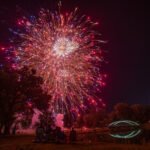 Fireworks explode in red, blue, and gold over a park at night, with spectators seated under trees watching near a lit bridge.