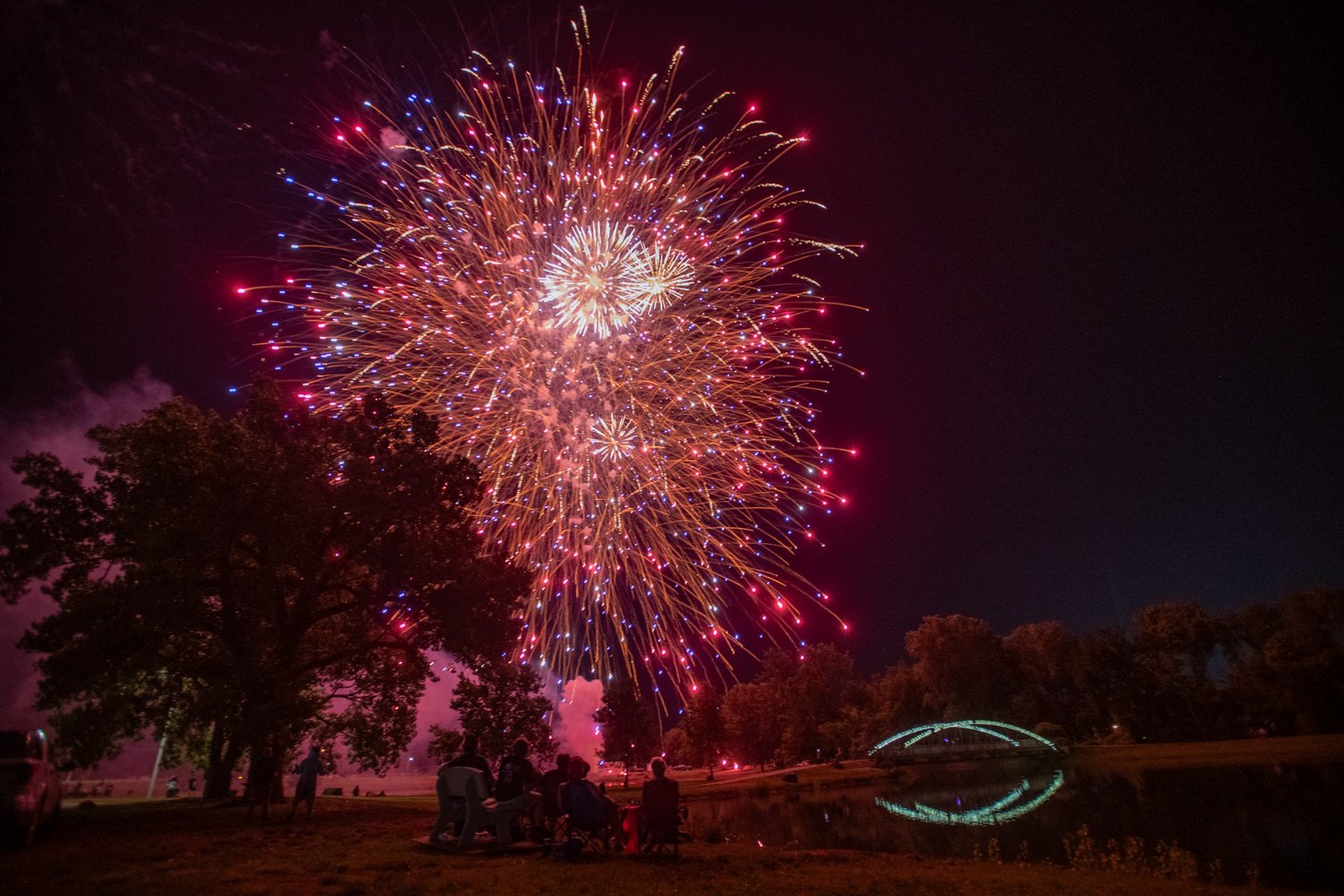 Fireworks explode in red, blue, and gold over a park at night, with spectators seated under trees watching near a lit bridge.