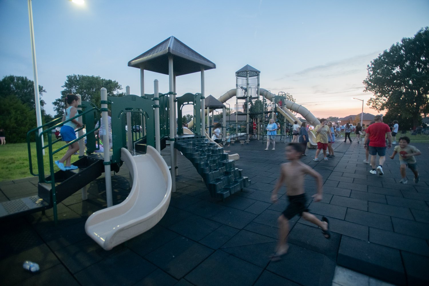Children run and play on a large outdoor playground at dusk, with slides, climbing bridges, and rubberized flooring in a park setting.