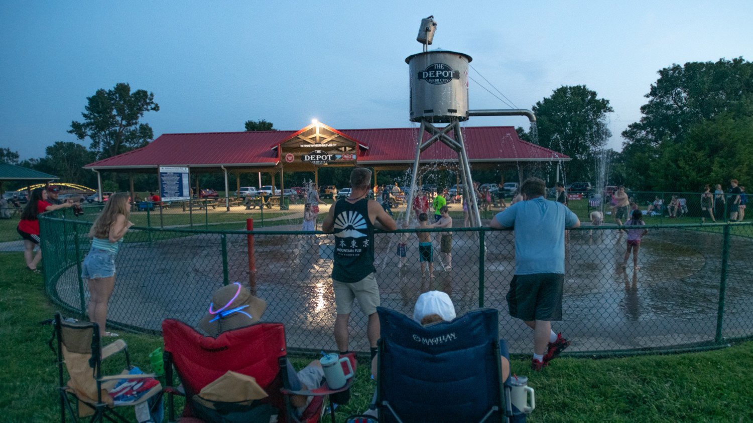 People watch children play in a sprinkling water area near a red-roofed pavilion at The Depot, fenced by a chain-link fence as a crowd gathers nearby.
