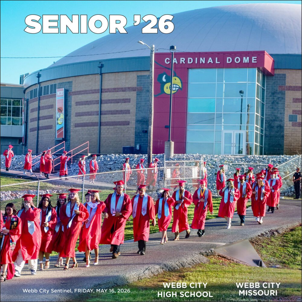 Graduating seniors in red caps and gowns walk along a fence-lined path outside Cardinal Dome, with 'SENIOR '26' across the sky.