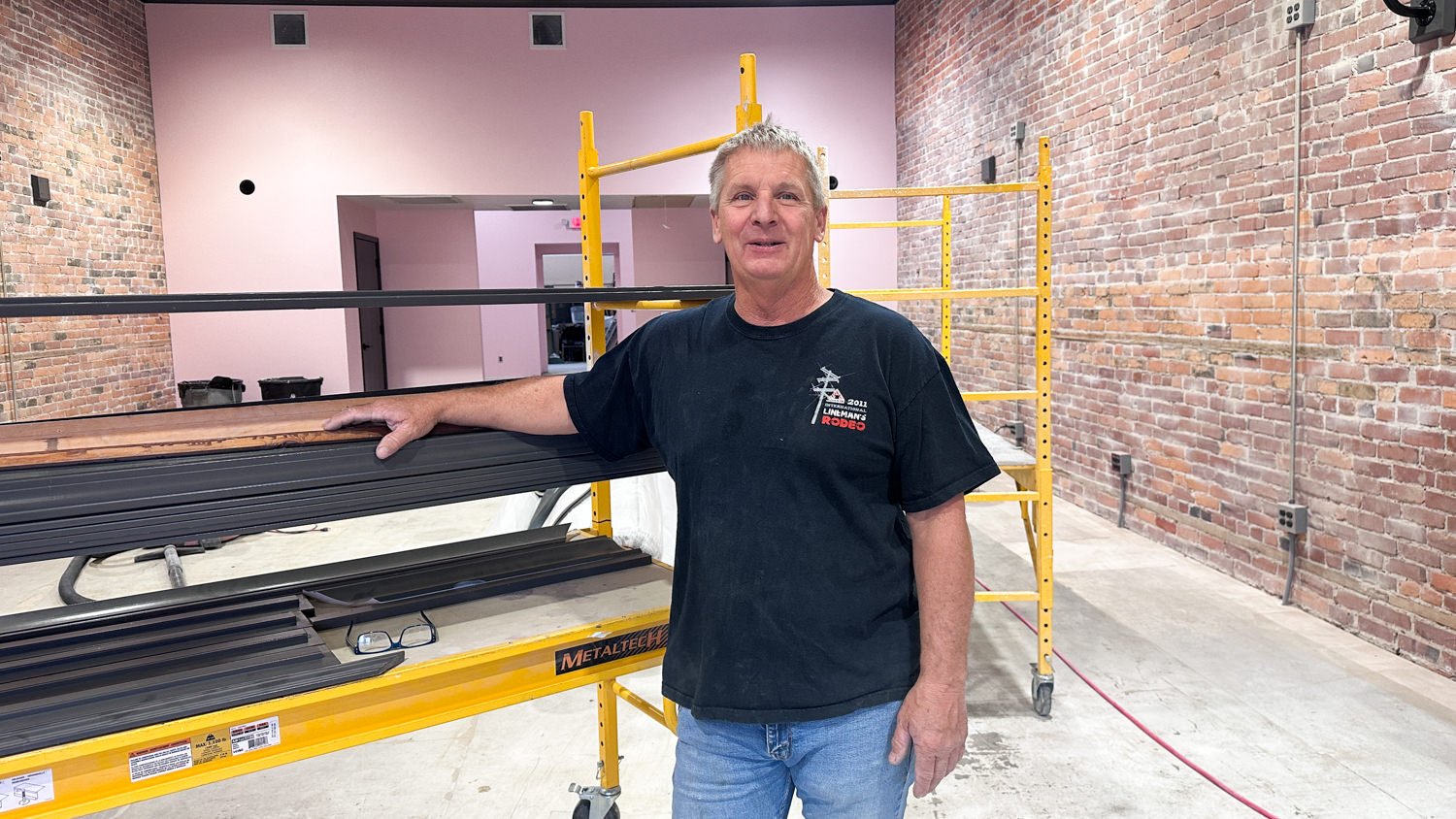 Man in a black T-shirt leaning on a stack of metal bars in a workshop with exposed brick walls and yellow scaffolding nearby.