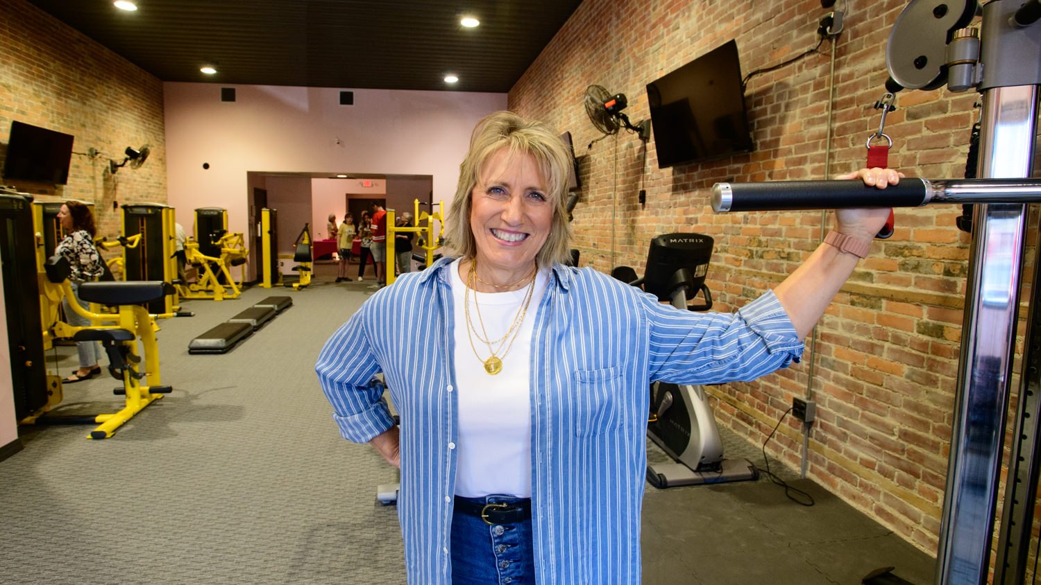 Smiling woman in a blue striped shirt stands in a brick-walled gym, resting her hand on a barbell rack in the foreground.