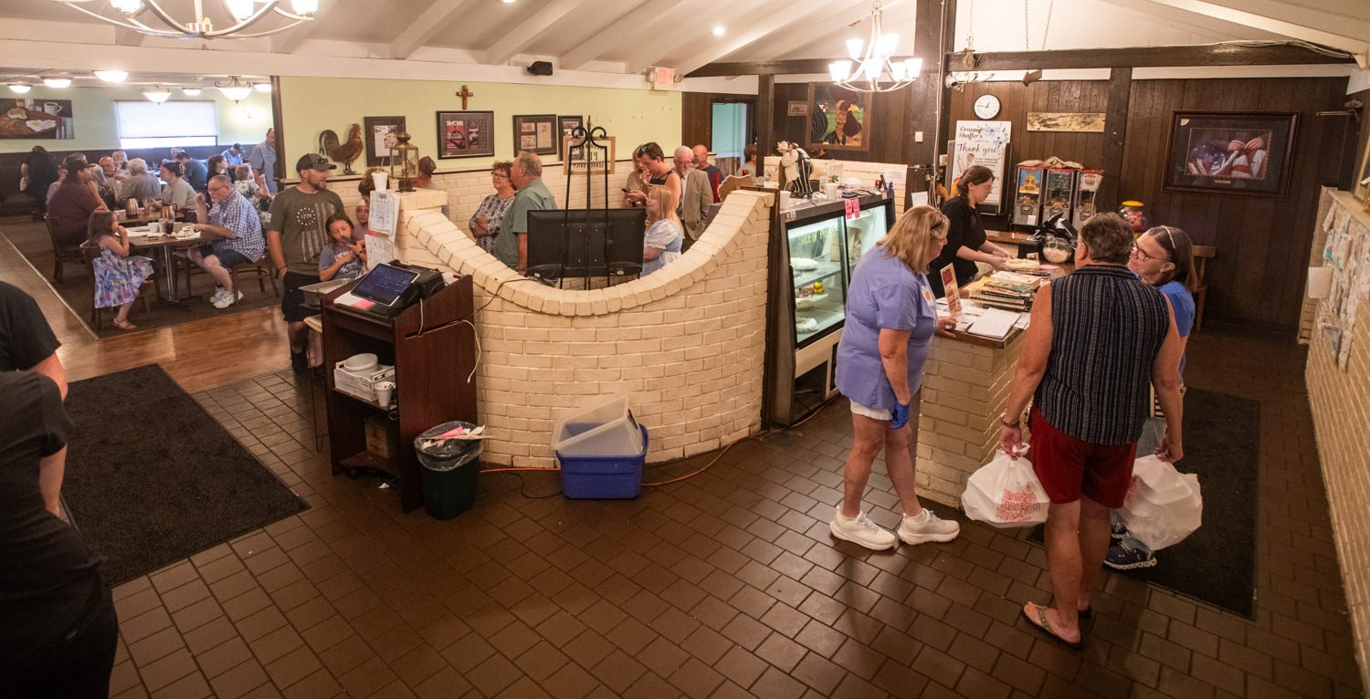 Group of people line up at a curved brick service counter in a busy dining hall; diners sit and eat in the background.