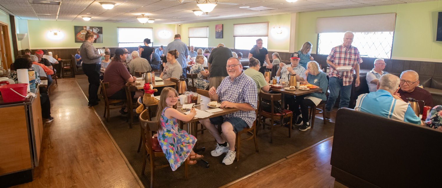 A busy diners hall with green walls and windows, where many people of different ages sit at wooden tables enjoying meals; in the foreground a smiling man in a checkered shirt sits with a young girl at a table.