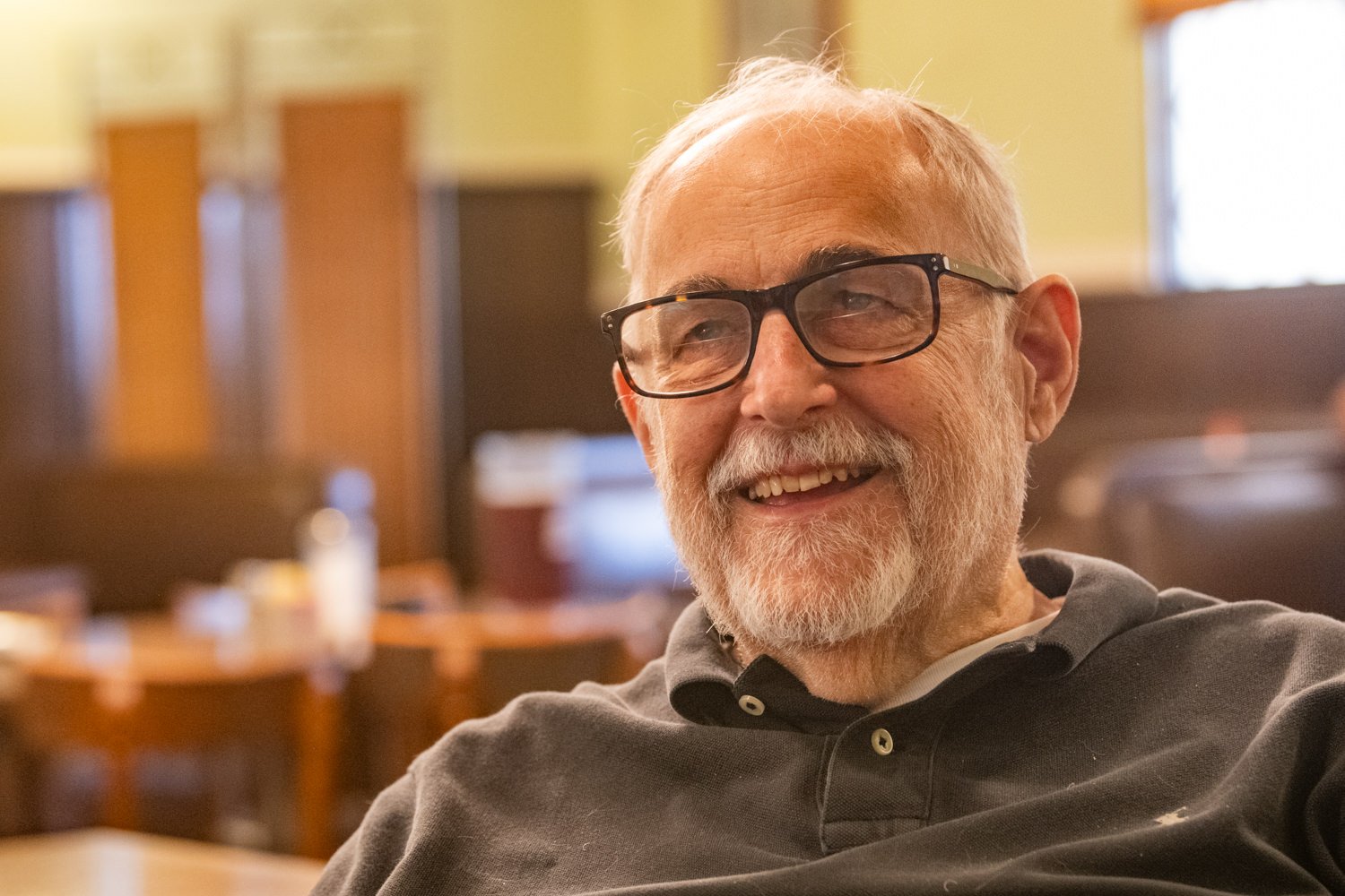 Smiling elderly man with white beard and black glasses in a cozy cafe setting.