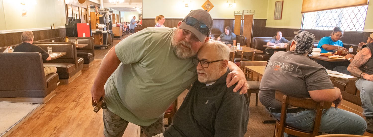 Two men share a friendly hug in a casual diner; one stands leaning over, wearing a green shirt and cap while the other sits in glasses and a dark polo; patrons in booths in background.