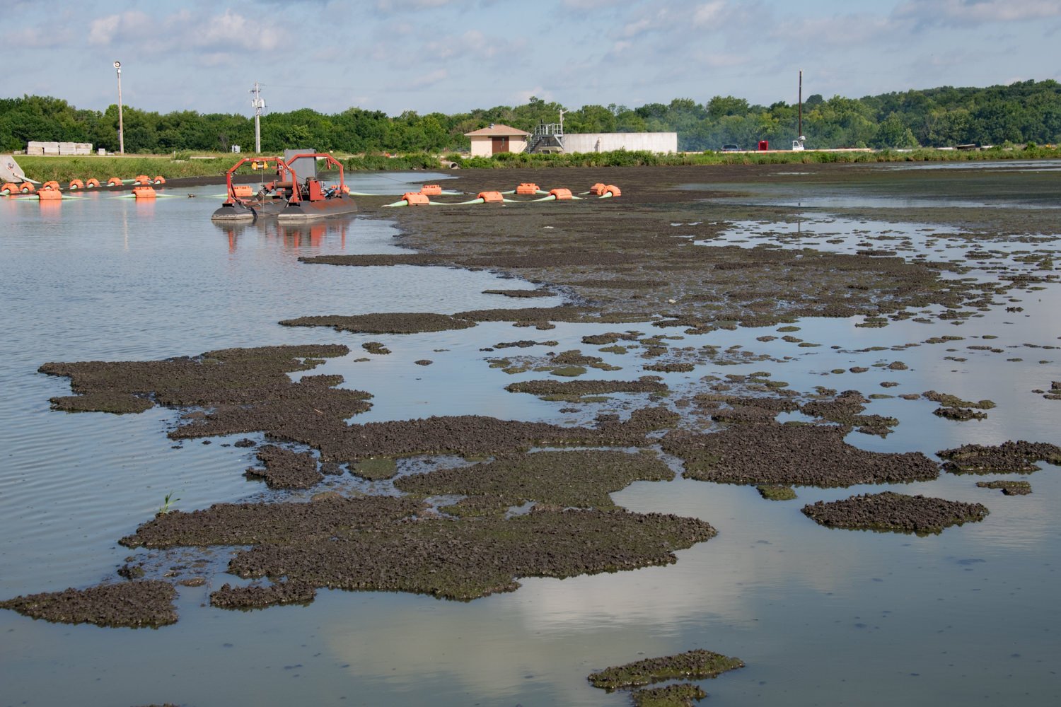 Mudflats and shallow water with orange buoys and a red dredging barge near shore, trees in the background.