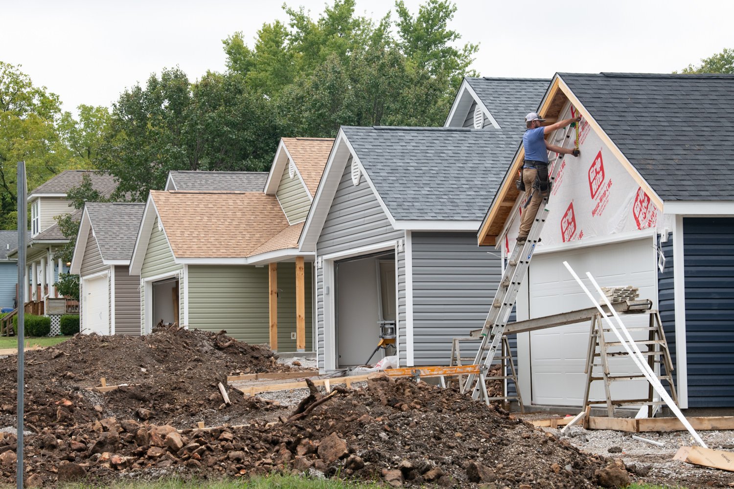 Construction worker on a ladder installing roof siding on a gray house; dirt piles and building materials in the yard.