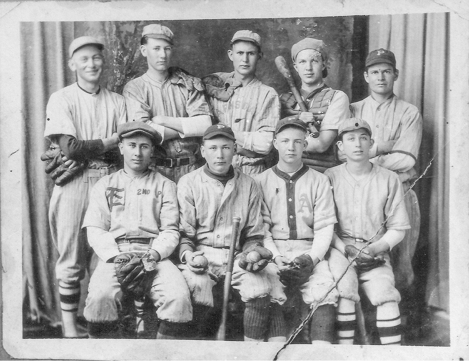A group of nine young baseball players in vintage uniforms posing for a studio team portrait, some holding gloves and bats, with one wearing catcher’s gear in the back row.