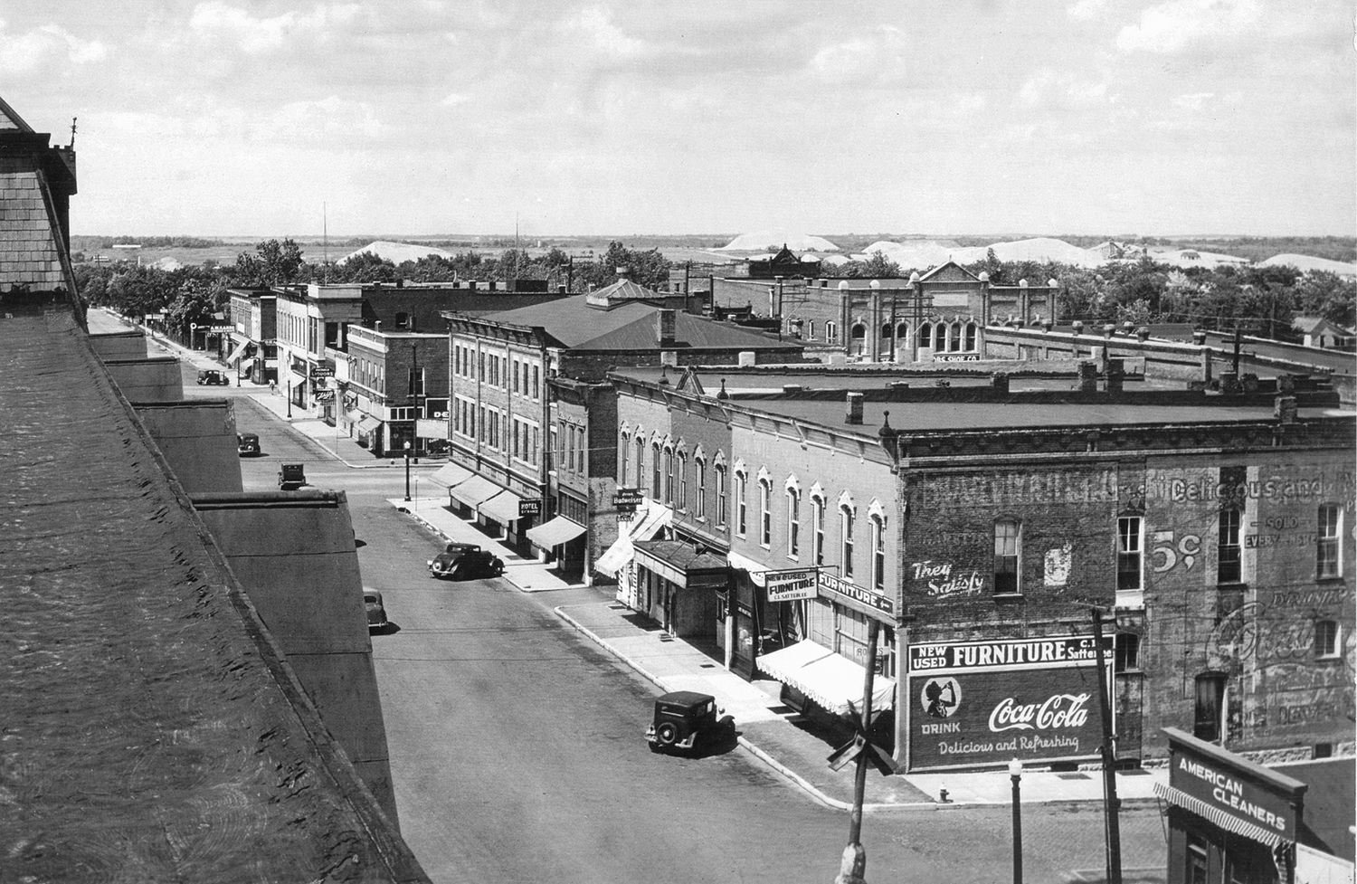 A black-and-white aerial view of a small-town main street with brick storefronts, vintage cars, and awnings along the sidewalk.
