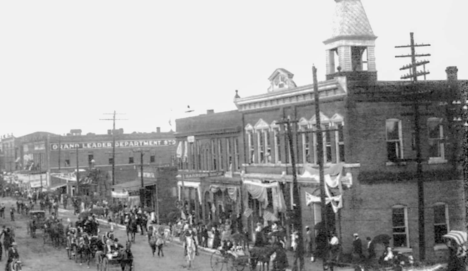 Historic street scene with a decorated brick storefront and banners, a crowd gathered as people and horse-drawn carriages parade down the wide street.