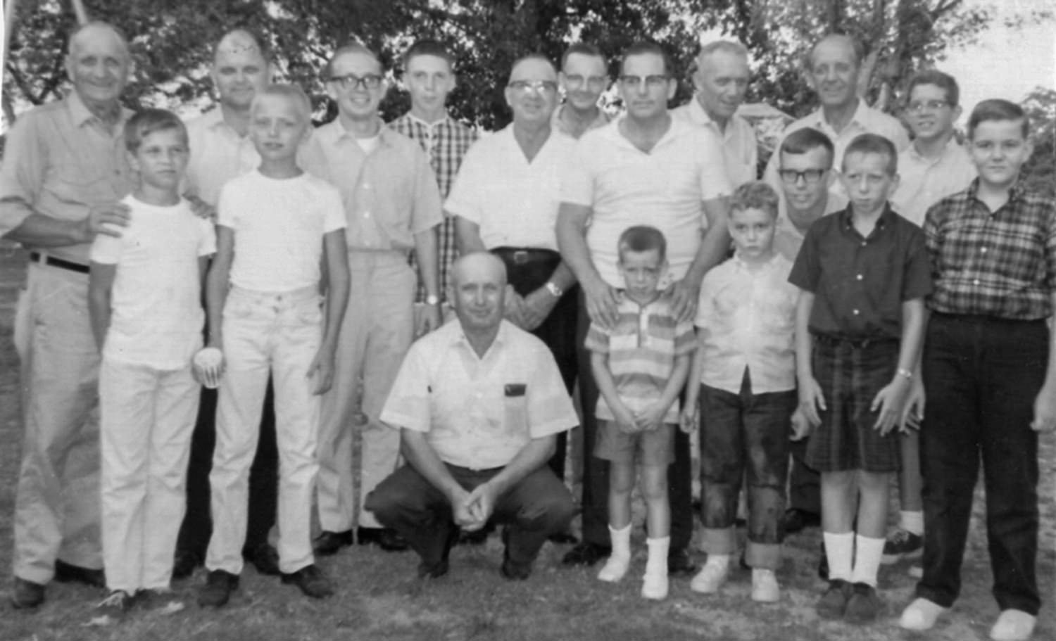 Group of men and boys posing outdoors for a black-and-white photo, several adults standing behind younger boys in a yard or park setting, daytime.