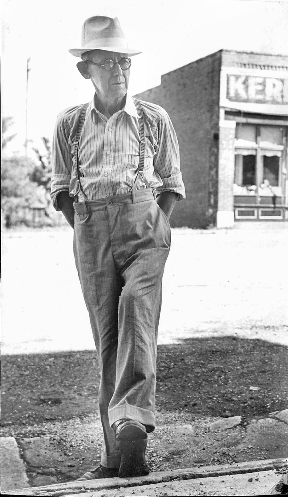 Older man wearing a fedora, striped shirt, suspenders, and high-waisted pants walking along a street past a brick storefront.
