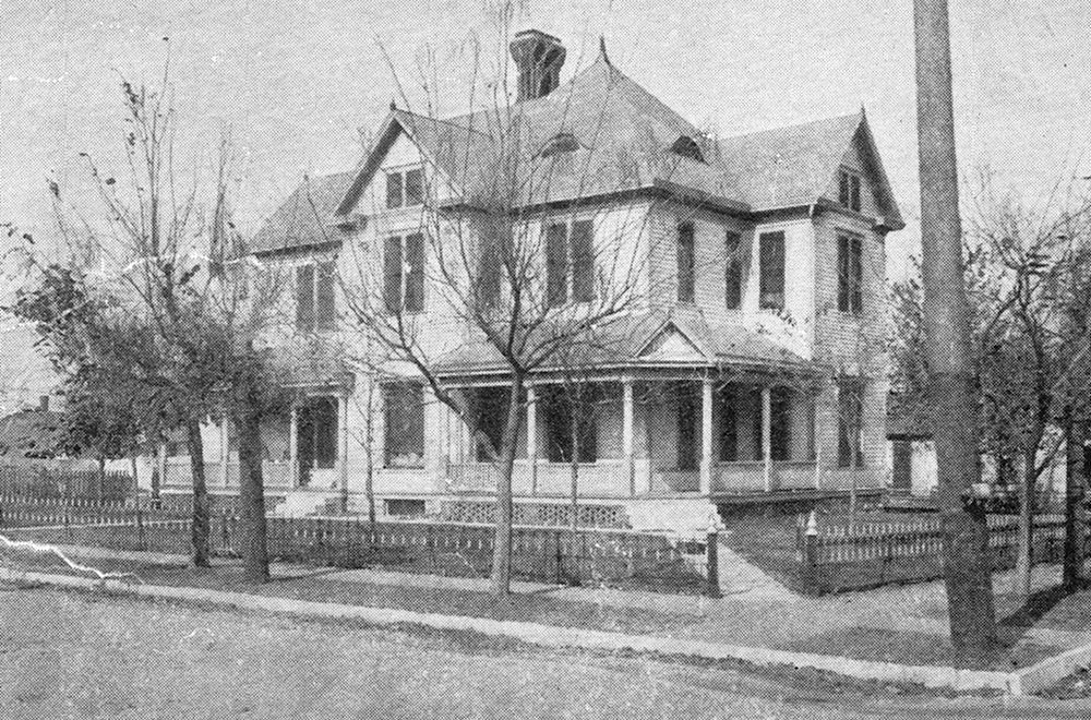 Two-story wooden house with a wraparound porch, bare trees in front, and a picket fence along a quiet street, conveying a historic feel.