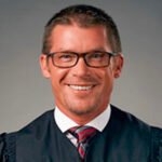 Smiling man in a dark judicial/academic robe with glasses, posed for a portrait.