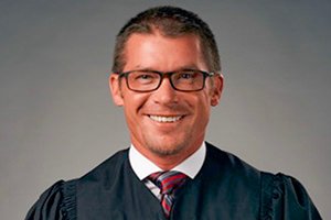 Smiling man in a dark judicial/academic robe with glasses, posed for a portrait.