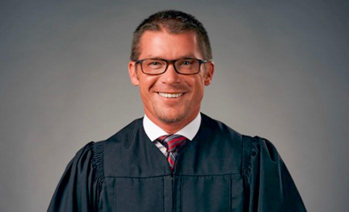 Smiling man in a black graduation gown and glasses, photographed against a neutral backdrop.