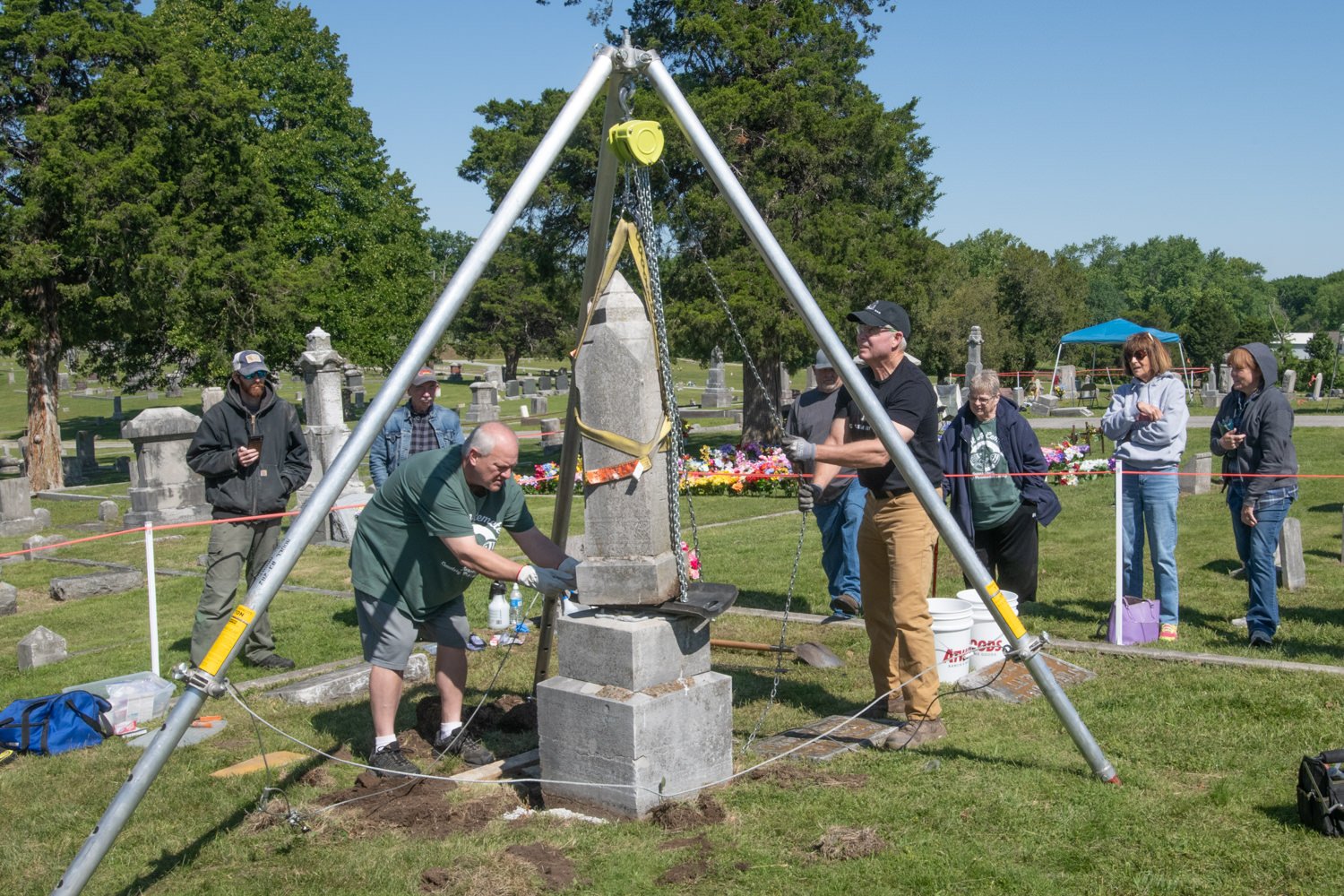 Volunteers install a stone obelisk on a pedestal in a cemetery using a tripod hoist, while onlookers stand nearby behind a cordon.